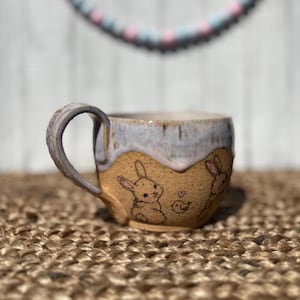 May include: A handmade ceramic mug with a light brown base and a light blue rim. The mug features illustrations of bunnies and a bird. The handle is a matching light blue color. The mug is sitting on a woven surface.