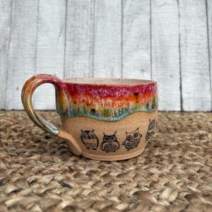May include: A handmade ceramic mug with a colorful glazed rim in shades of red, orange, yellow, and blue. The lower portion of the mug is tan with black owl illustrations. The handle is a matching color with the rim.