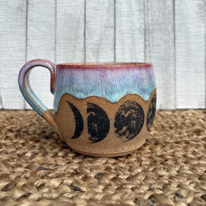 May include: A ceramic mug with a brown base and a light blue and purple rim. The mug features black moon phase designs around the body. The handle is a matching light blue and brown color.