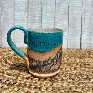 May include: A ceramic mug with a turquoise blue top and handle, and a tan base with a black mountain range design. The mug has a speckled interior and sits on a woven surface.