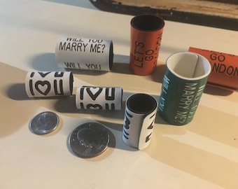 May include: A set of six white, black, green, and orange plastic rings with different messages printed on them. The messages include "Will You Marry Me?", "Let's Go", "I Love You", and "Go London".