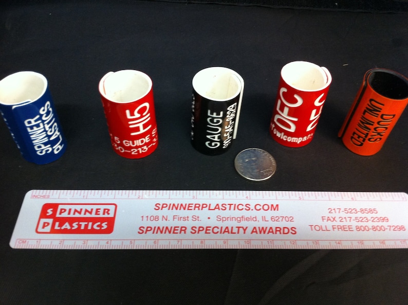 May include: Five colorful plastic cylinder-shaped labels with white text. The labels are red, black, orange, blue, and red. The text on the labels reads "Spinner Plastics", "H15 5 Guide 10-213", "Gauge 010-545-8629", "OFC ww/compac", and "Ducks Unlimited".