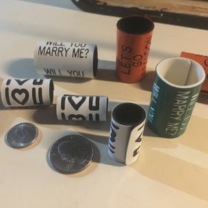 May include: A set of six white, black, green, and orange plastic rings with different messages printed on them. The messages include "Will You Marry Me?", "Let's Go", "I Love You", and "Go London".