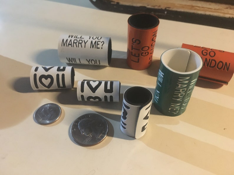 May include: A set of six white, black, green, and orange plastic rings with different messages printed on them. The messages include "Will You Marry Me?", "Let's Go", "I Love You", and "Go London".
