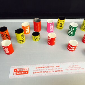 May include: A collection of colorful plastic rings with various text and numbers printed on them. The rings are used for identification and are made by Spinner Plastics.