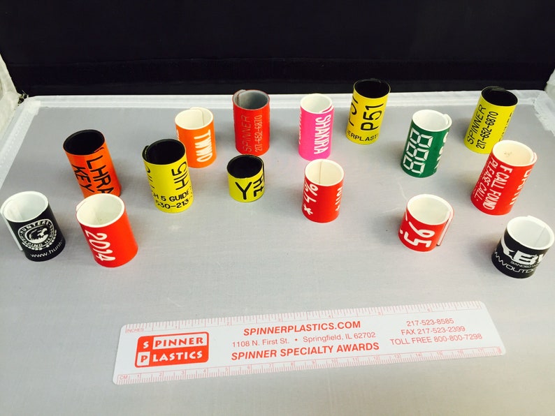 May include: A collection of colorful plastic rings with various text and numbers printed on them. The rings are used for identification and are made by Spinner Plastics.