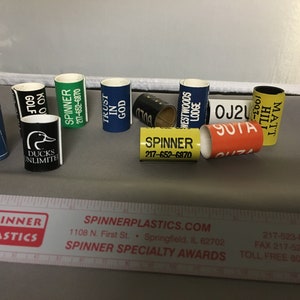 May include: A collection of colorful plastic award rings with various text and logos, including "Ducks Unlimited", "Spinner", "Trust in God", "Westwoods Lodge", "Matt Hill", "OJ2U", and "SUTA".