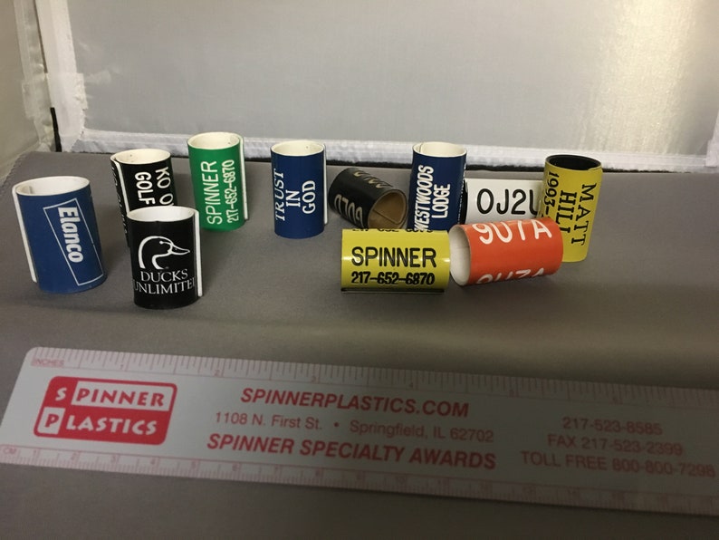 May include: A collection of colorful plastic award rings with various text and logos, including "Ducks Unlimited", "Spinner", "Trust in God", "Westwoods Lodge", "Matt Hill", "OJ2U", and "SUTA".