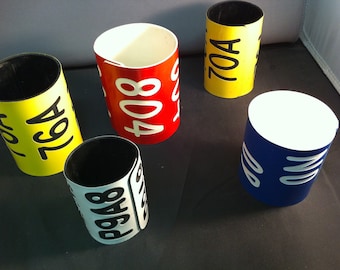 May include: A collection of colorful, cylindrical containers. The containers are painted in various colors, including yellow, red, white, and blue. Each container has white text with numbers and letters. The containers are arranged on a dark surface.