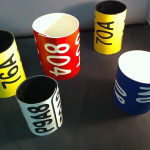 May include: Five colorful cylindrical containers with numbers printed on them. The containers are yellow, red, blue, and white with black numbers. The numbers are 76A, 408, 70A, 848, and 20.