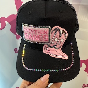 Könnte beinhalten: Schwarze Trucker-Cap mit Mesh-Rückseite, mit einem rosa Aufnäher mit der Aufschrift "WESTERN BABES" und einem rosa Cowboy-Stiefel-Applikation. Der Schirm ist mit schillernden Strasssteinen besetzt.
