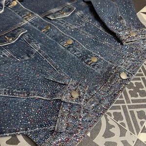 Könnte beinhalten: Dunkelblaue Jeansjacke mit schimmernden Strasssteinen verziert. Die Jacke hat klassische Details wie Knopfverschlüsse, Brusttaschen und einen Kragen. Die Manschetten und Kanten sind stark mit Kristallen verziert, was einen glamourösen Look erzeugt.