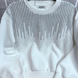 Może przedstawiać: Biała bluza z długimi rękawami z wzorem kaskadowych srebrnych kryształków. Bluza ma okrągły dekolt i ściągacze przy mankietach. Na metce widnieje nazwa marki "MISSKISS".