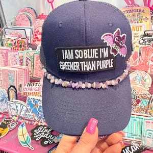 Könnte beinhalten: Marineblaue Trucker-Cap mit dem Aufnäher "I AM SO BLUE I'M GREENER THAN PURPLE". Sie hat eine lila Fledermaus und einen Amethyst-Perlenbesatz. Die Kappe hat einen Mesh-Rücken und einen gebogenen Schirm.