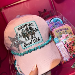 Peut inclure: Casquette de baseball rose avec une bordure de pierres turquoise et une étoile argentée. La casquette porte un écusson avec les mots "Cowgirl Social Club" et un graphisme de cowgirls. La casquette est dans une boîte rose avec d'autres accessoires.