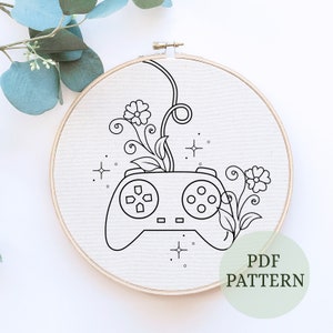 May include: A black and white line drawing of a video game controller with flowers growing out of the top. The image is on a white background and has the text "PDF PATTERN" in the bottom right corner.