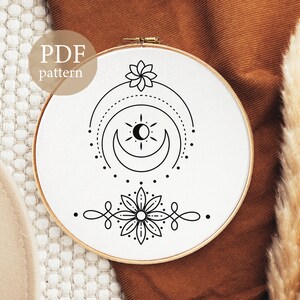 May include: A black and white embroidery pattern featuring a crescent moon, sun, and two stylised flowers. The pattern is displayed on a white embroidery hoop.