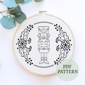 May include: A black and white Nutcracker design on a cream-colored fabric, framed in a wooden embroidery hoop. The design is surrounded by a floral and leaf border. The text "PDF PATTERN" is in a green circle.