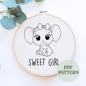 以下が含まれることがあります： かわいい象がリボンをつけている、白黒の線画刺繍パターン。象の下に「Sweet Girl」という文字があります。パターンは白い背景にあり、木製の刺繍枠に入っています。