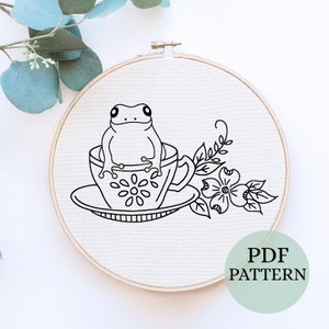 Puede incluir: Patrón de bordado en línea de arte en blanco y negro de una rana sentada en una taza de té con un platillo y flores. El patrón está etiquetado como "PDF PATTERN".