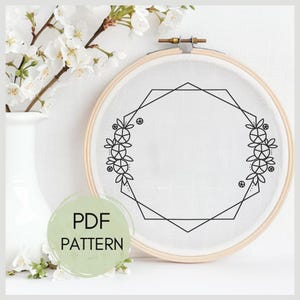 May include: A black and white embroidery pattern featuring a geometric octagon frame with floral accents. The pattern is displayed on a white fabric in a wooden embroidery hoop.