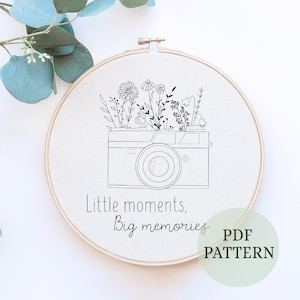 May include: Black and white embroidery pattern of a camera with flowers growing out of it. The text "Little moments, Big memories" is below the camera. The pattern is on a white background and is labeled "PDF PATTERN".