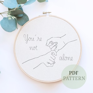 Puede incluir: Un dibujo en blanco y negro de dos manos que se agarran los meñiques, con el texto "You're not alone" sobre las manos. El dibujo está sobre un fondo blanco y está enmarcado en un aro de madera. El texto "PDF PATTERN" está escrito debajo del dibujo.