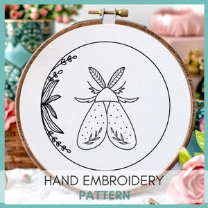 Peut inclure: Motif de broderie noir et blanc d'un papillon de nuit avec des d&eacute;tails floraux sur un tissu blanc dans un cerceau de broderie en bois. Le cerceau a un diam&egrave;tre de 15 cm. Le texte "HAND EMBROIDERY PATTERN" est imprim&eacute; sous le cerceau.