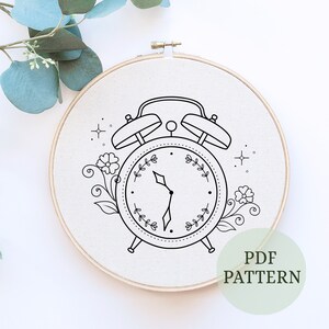 May include: A black and white line drawing of an alarm clock with floral accents. The clock face has a simple design with two hands and a wreath around the edge. The text "PDF PATTERN" is visible in the lower right corner of the image.