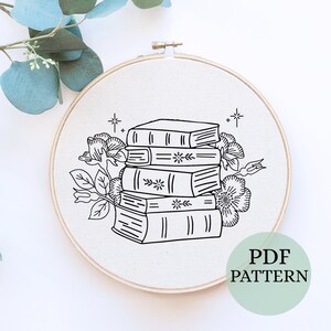 May include: A black and white line drawing of a stack of books with floral embellishments. The image is in a circular embroidery hoop. The text "PDF PATTERN" is in the lower right corner of the image.