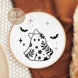 Op de afbeelding: Een zwart-witte borduurring met een spookontwerp, versierd met bloemmotieven, een strik en vleermuizen. De tekst "PDF pattern" is zichtbaar. Het ontwerp staat op een witte achtergrond in een houten ring.
