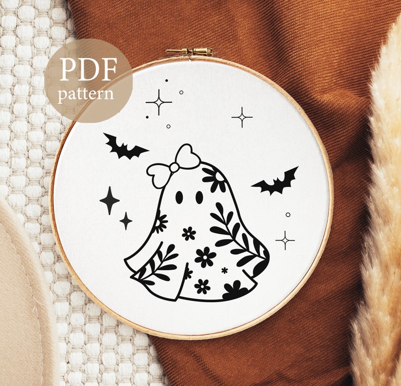 Op de afbeelding: Een zwart-witte borduurring met een spookontwerp, versierd met bloemmotieven, een strik en vleermuizen. De tekst "PDF pattern" is zichtbaar. Het ontwerp staat op een witte achtergrond in een houten ring.