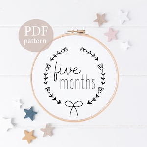 May include: A white embroidery hoop with a black floral wreath and the text "five months" inside. The hoop is on a white wooden background with small wooden stars scattered around it.