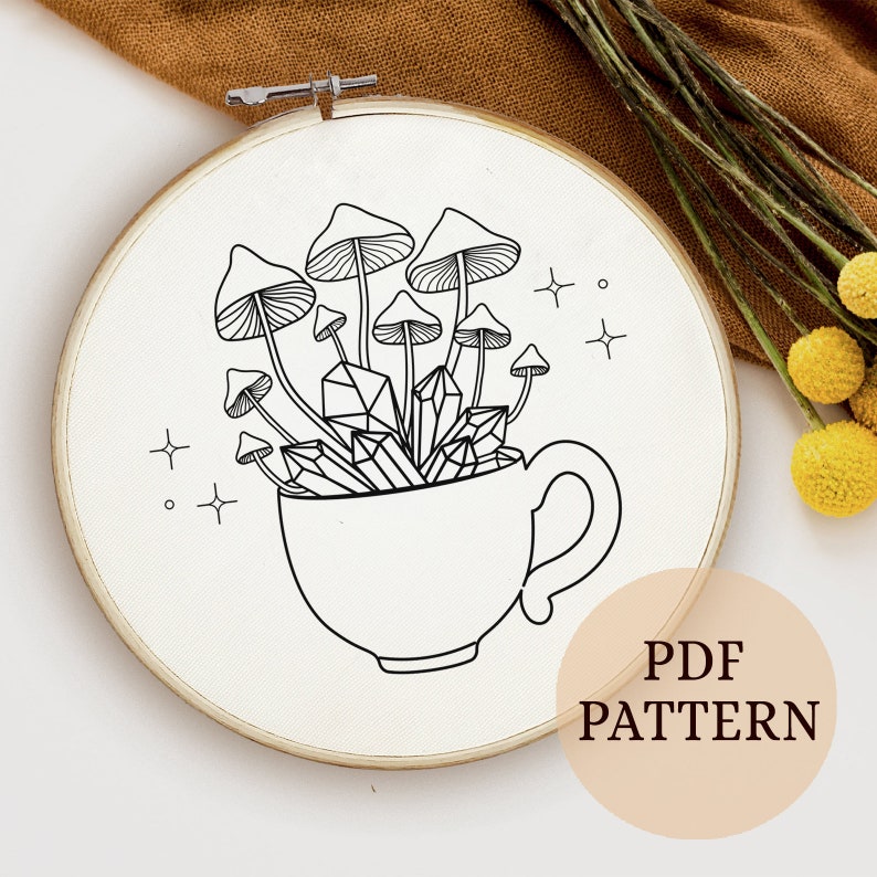 May include: A black and white line drawing of a teacup filled with mushrooms and crystals. The teacup has a handle and is surrounded by stars. The image is on a white background and is framed in a wooden embroidery hoop.