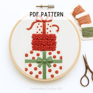 Op de afbeelding: Een borduurring met een gestikt ontwerp van drie gestapelde cadeau dozen. De bovenste doos heeft een rode strik, de middelste doos is bedekt met rode bloemen en de onderste doos heeft rode stippen en een groene strik. De tekst "PDF PATTERN" staat bovenaan.