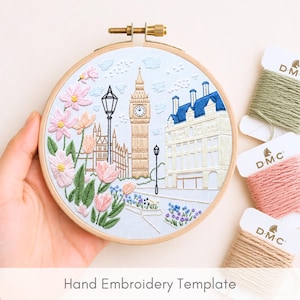 May include: A round wooden embroidery hoop containing fabric with a stitched scene of Big Ben, a street lamp, and a building, surrounded by pink and green floral embroidery. Three spools of DMC embroidery floss in green, pink, and tan are included.