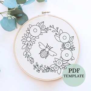 May include: A black and white embroidery pattern of a bee surrounded by a wreath of flowers. The pattern is on a white fabric in a wooden embroidery hoop. The text "PDF TEMPLATE" is in the lower right corner of the image.
