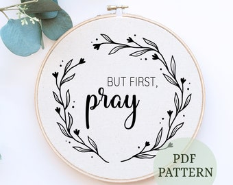 May include: A white embroidery hoop with a black floral wreath and the text "But first, pray". The hoop is on a white background with a green plant in the top left corner. The text "PDF PATTERN" is in a green circle in the bottom right corner.