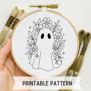 May include: A white embroidery hoop with a black and white ghost design surrounded by flowers and leaves. The text "PRINTABLE PATTERN" is displayed on a white banner. Several spools of green and gold embroidery floss are visible.