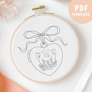 May include: A black and white line drawing of a cat's face inside a heart-shaped locket. The locket is attached to a ribbon with a bow. This is a printable PDF template for embroidery.