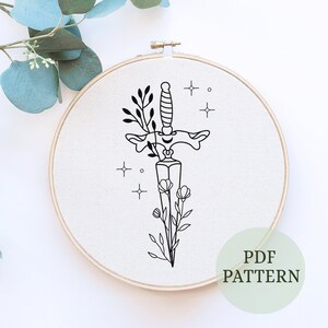 May include: A black and white embroidery pattern of a dagger with flowers and stars. The pattern is on a white fabric in a wooden embroidery hoop. The text "PDF PATTERN" is in the lower right corner of the image.