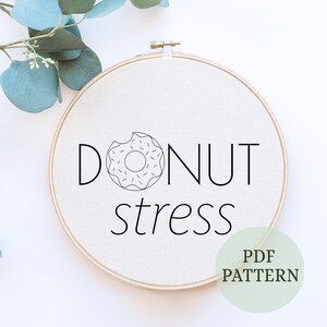 May include: A white embroidery hoop with a black outline of a donut and the text "Donut stress" in black. The hoop is on a white background with a green leaf in the top left corner. The text "PDF PATTERN" is in a green circle in the bottom right corner.