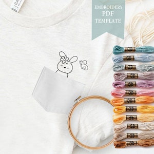 May include: A white t-shirt with a pocket and an embroidered bunny and butterfly design. The t-shirt is accompanied by a wooden embroidery hoop and a variety of embroidery floss in various pastel colors. The text "EMBROIDERY PDF TEMPLATE" is displayed in a light blue box.