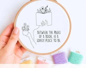 Könnte beinhalten: Ein Handstickvorlage in einem Holzrahmen. Das Design zeigt eine Person mit einem Buch als Kopf, die einen kleinen Topf mit Blumen hält. Der Text lautet: „ZWISCHEN DEN SEITEN EINES BUCHES IST ES EIN SCHÖNER ORT ZU SEIN.“ Stickgarn in Gelb, Rosa, Lila und Grün.