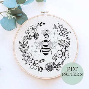 May include: Black and white embroidery pattern featuring two bees and a floral wreath. The pattern is displayed on a white fabric stretched over a wooden embroidery hoop. The text "PDF PATTERN" is visible in the lower right corner of the image.