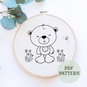 May include: Black and white line art embroidery pattern of a smiling bear standing in a field of flowers with a bee flying above. The pattern is on a white fabric in a wooden embroidery hoop. The text "PDF PATTERN" is written below the hoop.