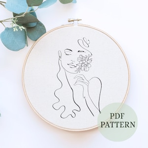 May include: A black and white line drawing of a woman's face with flowers on one side. The drawing is in a wooden embroidery hoop. The text "PDF PATTERN" is written below the drawing.