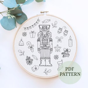 May include: Black and white line art illustration of a nutcracker with Christmas decorations. The illustration includes a nutcracker, candy canes, mittens, a star, a gift box, a bell, and a candle. The text "PDF PATTERN" is in the lower right corner of the image.