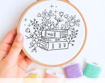 May include: A hand embroidery template featuring a floral design with a stack of books. The design is in black ink on white fabric, set in a wooden embroidery hoop. The text "HAND EMBROIDERY TEMPLATE" is at the bottom. Colorful embroidery floss is also visible.