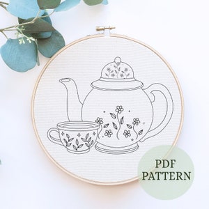 May include: A black and white line drawing of a teapot and teacup with floral designs. The image is on a white background and is framed in a wooden embroidery hoop. The text "PDF PATTERN" is written in green on a white background.